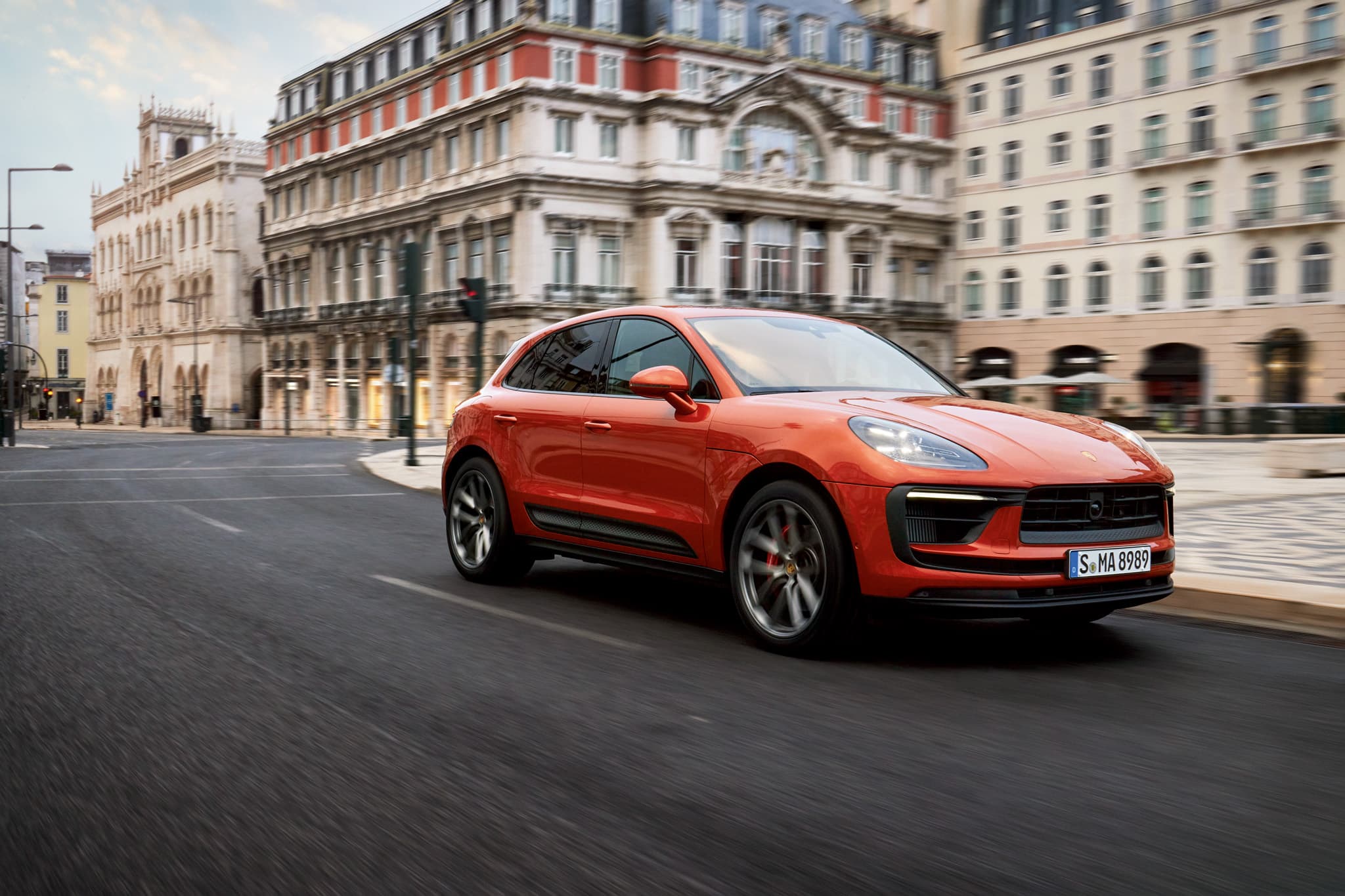 Porsche Macan S in Papaya Metallic drives through a city street with ornate, historic buildings in the background.